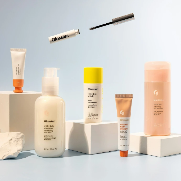 Assorted skincare products and a mascara wand arranged on white pedestals against blue